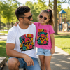A man and a young girl wearing matching graphic t-shirts in an outdoor park setting. The man wears a white t-shirt with a colorful 'ONE RAD DAD' design, and the girl wears a pink t-shirt with a matching 'RAD LIKE DAD' design.