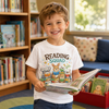 A young boy smiling in a library or bookroom, wearing a white graphic T-shirt that says 'READING SQUAD' with cartoon animals reading books. He is holding an open book.