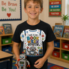 A young boy smiling in a classroom while wearing a black graphic t-shirt that reads 'THIS IS MY BOOK WEE costume!'. He is holding a small robot toy.