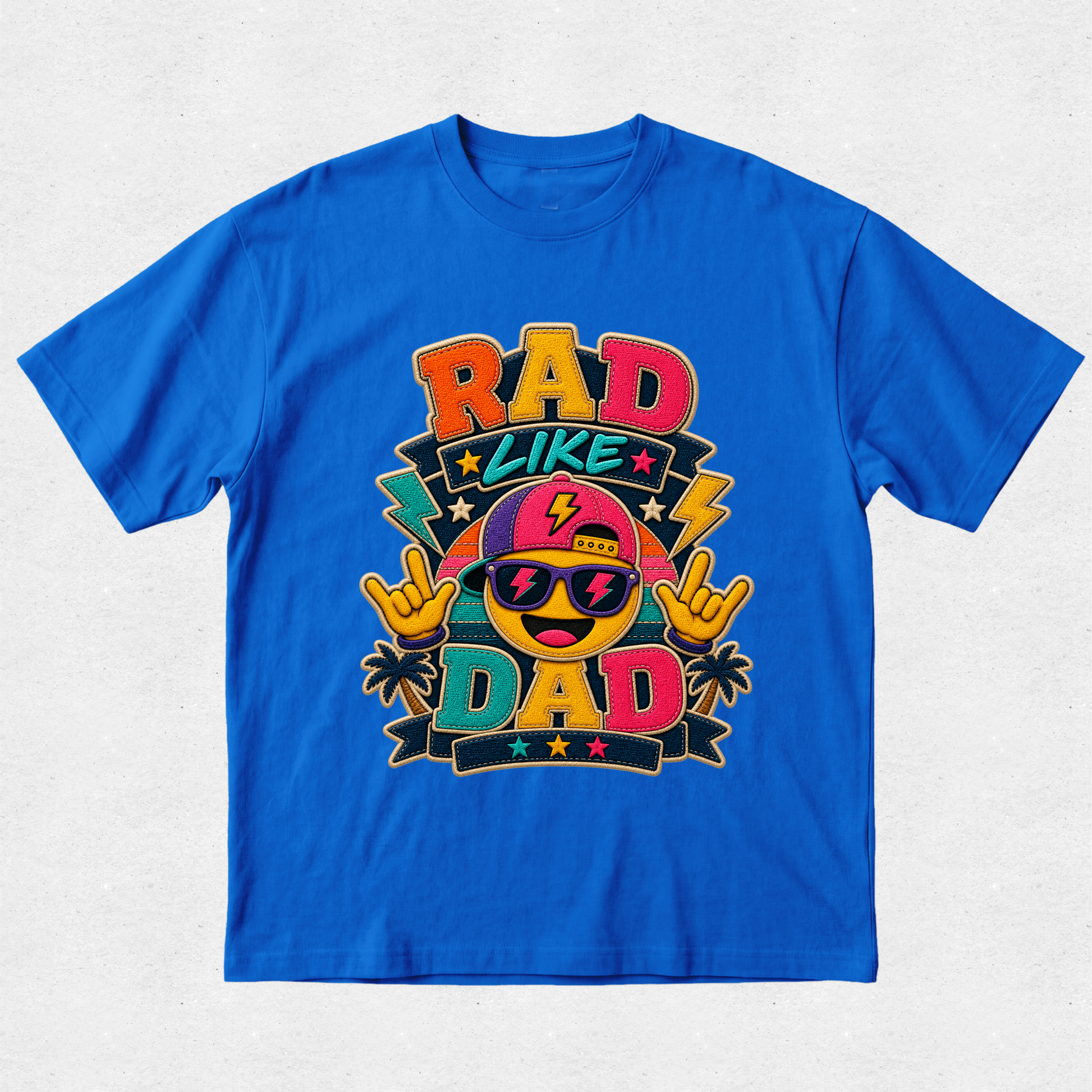 A bright blue short-sleeve t-shirt with a colorful graphic on the front that reads 'RAD LIKE DAD' with a cartoon emoji wearing sunglasses and a cap, set against a light gray textured background.