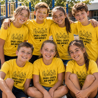 Six children smiling outdoors in matching bright yellow T-shirts with black text reading 'SMALL CHATS BIG IMPACT' and a small illustrated graphic above it. The background shows a playground with blue and yellow equipment.
