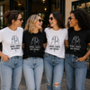Four women standing outdoors and smiling, each wearing a graphic T-shirt. Two shirts are white and two are black, with a matching dog illustration and the text 'SMALL CHATS BIG IMPACT.