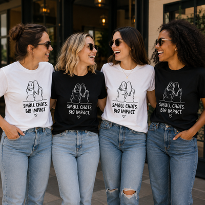 Four women standing outdoors and smiling, each wearing a graphic T-shirt. Two shirts are white and two are black, with a matching dog illustration and the text 'SMALL CHATS BIG IMPACT.