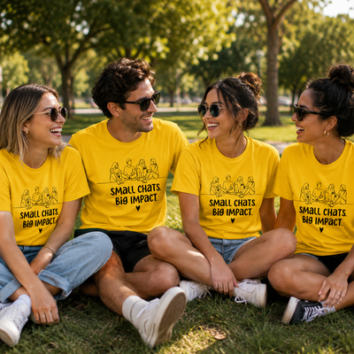 Four people sitting on grass in a park wearing matching bright yellow graphic T-shirts with the text 'SMALL CHATS. BIG IMPACT.