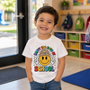 A young boy smiling in a classroom, wearing a white graphic t-shirt that reads 'Happy 100 Days of School' with a yellow smiley face wearing a beanie and peace signs.