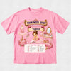 A pink short-sleeve graphic T-shirt laid flat on a light textured background. The shirt features a colorful ballet-themed 'Book Week Quest' print with the name 'Tamsin' on a banner, an illustration of a ballerina, a mannequin in a tutu, a vanity table, and a checklist at the bottom.