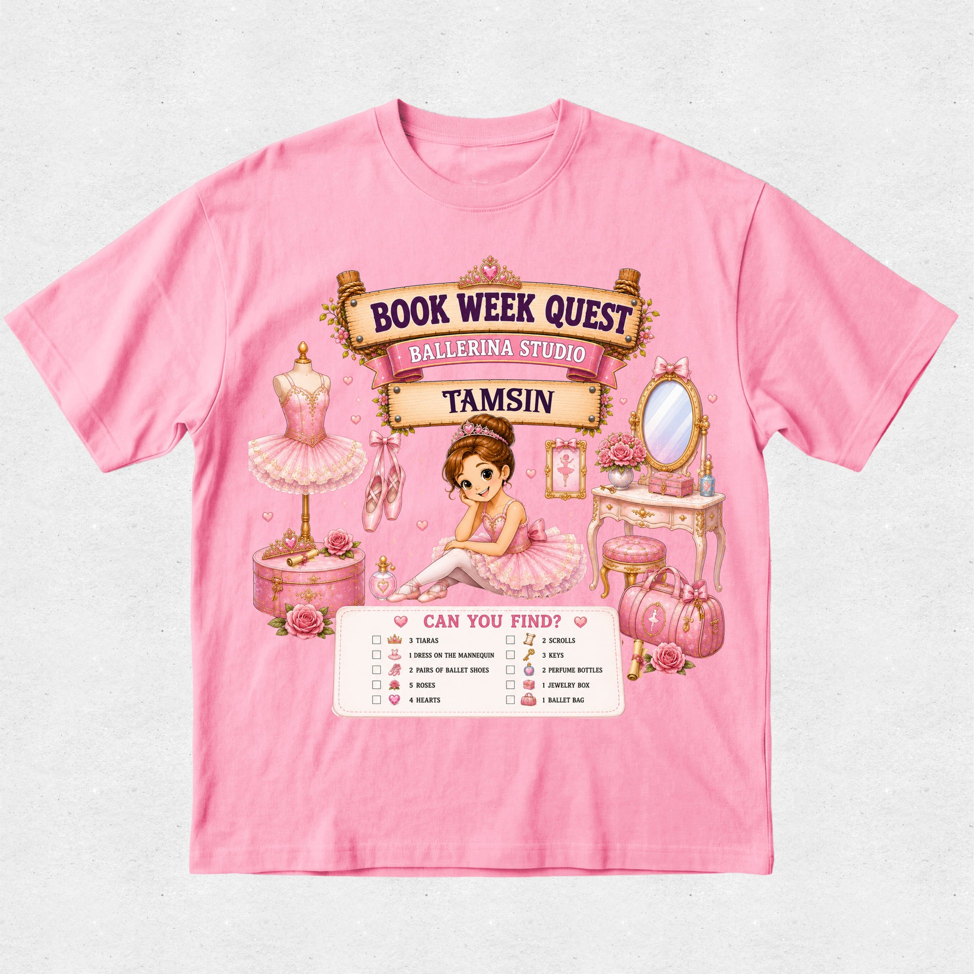 A pink short-sleeve graphic T-shirt laid flat on a light textured background. The shirt features a colorful ballet-themed 'Book Week Quest' print with the name 'Tamsin' on a banner, an illustration of a ballerina, a mannequin in a tutu, a vanity table, and a checklist at the bottom.