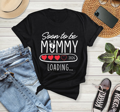 Black t-shirt with 'Soon to be Mummy 2026 Loading' text on a wooden surface with jeans, sneakers, and a phone.
