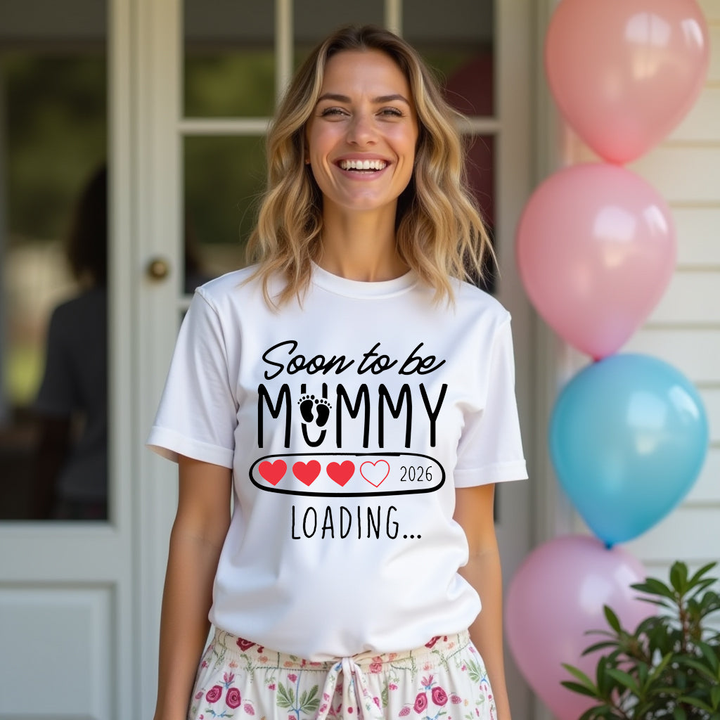 Woman wearing a white t-shirt with 'Soon to be Mummy Loading 2026' text, standing in front of balloons.