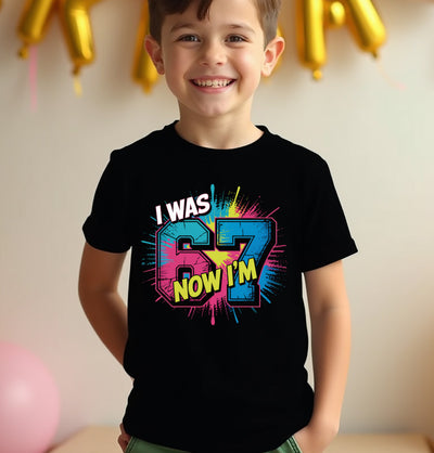 Child wearing a black t-shirt with colorful text 67, standing against a neutral background with balloons.