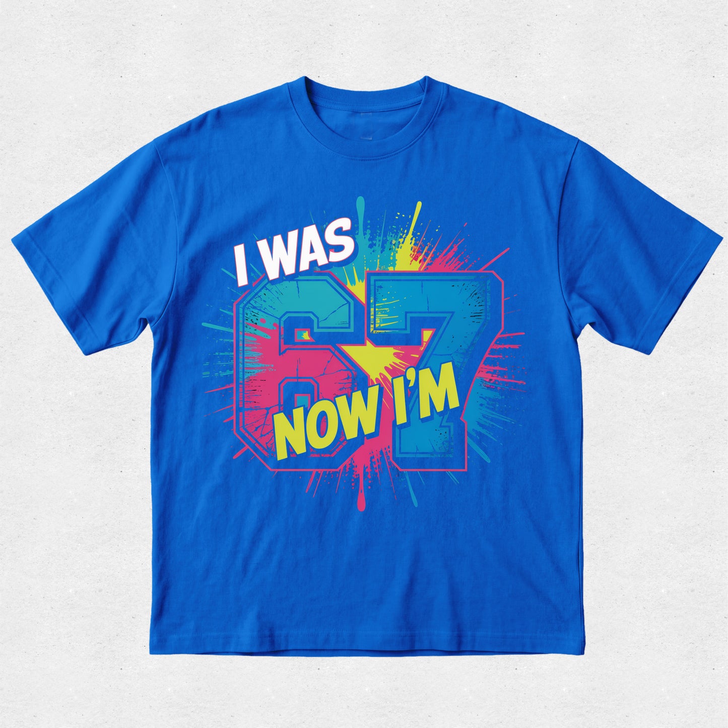 Blue t-shirt with colorful graphic text 'I WAS 67 NOW I'M' on a light gray background