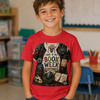 A young boy smiling in a classroom, wearing a red graphic t-shirt that reads 'THIS IS MY BOOK WEEK COSTUME' with a collage of books, skulls, and a cat.