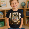 A young boy smiling in a classroom setting, wearing a navy blue graphic t-shirt with a vintage bookist costume print that reads 'THIS IS MY BOOK WEEK COSTUME!' and blue jeans.