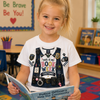 A young girl with blonde hair in a ponytail smiles while holding an open book in a classroom. She is wearing a white graphic t-shirt featuring a detailed police officer costume design with badges, a helmet, and the text 'THIS IS MY BOOK WEEK COSTUME!'.