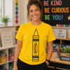 A smiling woman wearing a bright yellow graphic T-shirt with a large black crayon illustration and the word 'YELLOW' on the front, standing in a colorful classroom setting.