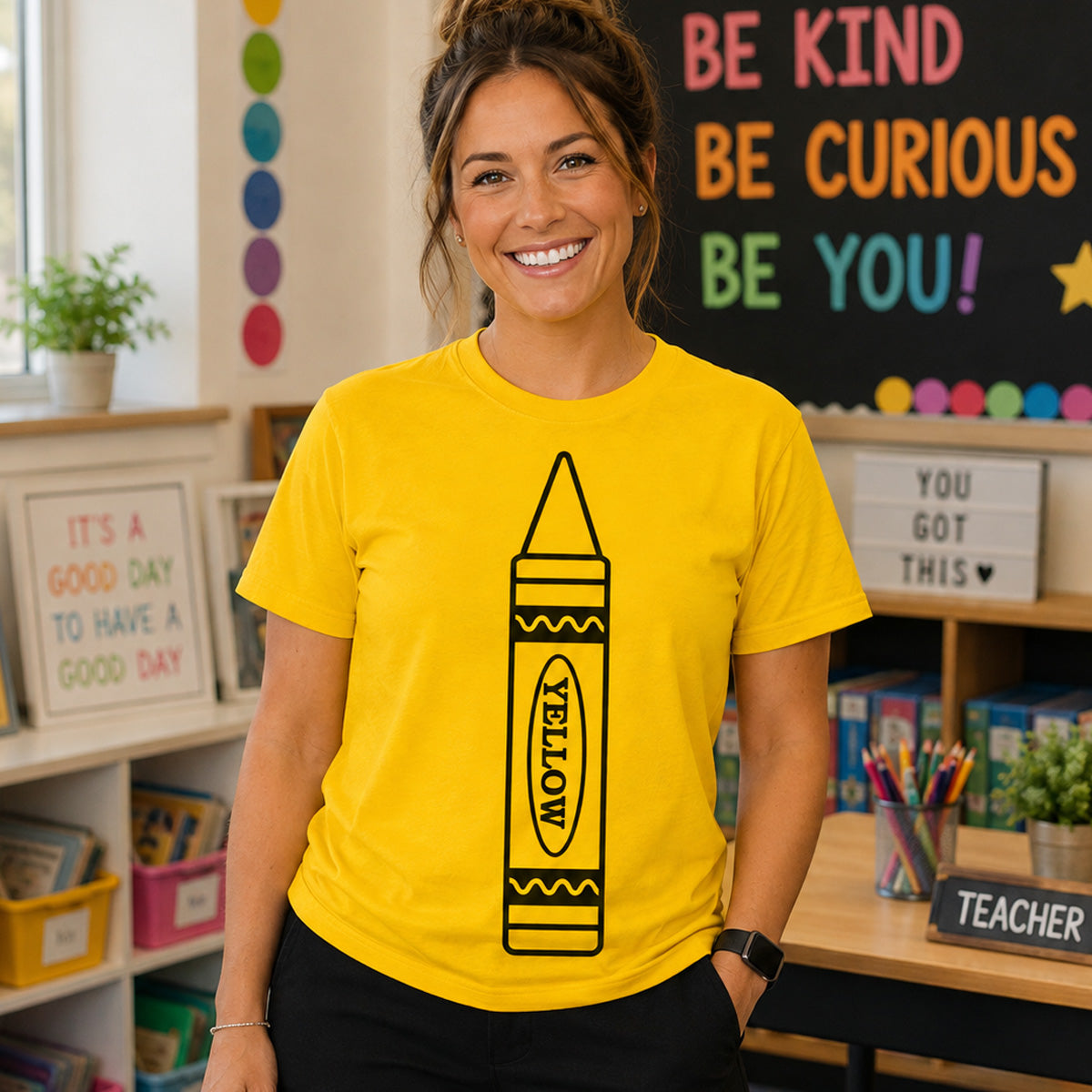 A smiling woman wearing a bright yellow graphic T-shirt with a large black crayon illustration and the word 'YELLOW' on the front, standing in a colorful classroom setting.