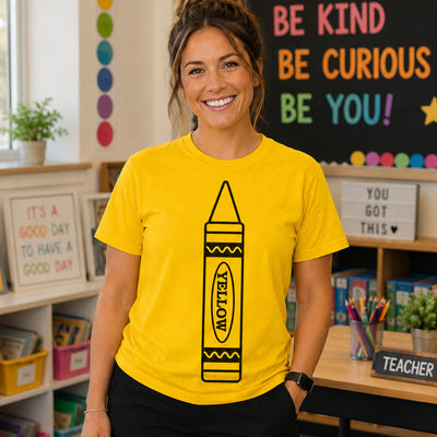 A smiling woman wearing a bright yellow graphic T-shirt with a large black crayon illustration and the word 'YELLOW' on the front, standing in a colorful classroom setting.