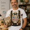 A smiling man in a book-themed costume stands in a classroom or library setting, wearing a white graphic T-shirt that reads 'THIS IS MY BOOK WEEK COSTUME!' with vintage explorer illustrations, and holding a book titled 'Great Explorers'.