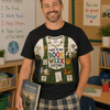 A smiling man stands in a classroom wearing a black graphic T-shirt with a colorful book-themed costume print on the front, paired with plaid shorts and holding a book.
