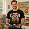 A smiling man in a classroom setting wearing a black graphic t-shirt with a pirate book week costume design, holding an open book. The background shows bookshelves and educational posters.