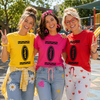 Three women standing together outdoors in front of a playground, each wearing a bright crayon-themed t-shirt in yellow, pink, and red, paired with jeans and patterned drawstrings.