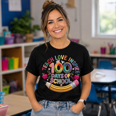 A woman smiling in a classroom setting, wearing a black graphic t-shirt that reads 'TEACH LOVE INSPIRE 100 DAYS OF SCHOOL' in colorful rainbow lettering with a pencil and apple design.