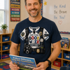 A smiling man in a classroom wearing a navy blue graphic t-shirt with a police officer costume design and holding a children's book titled 'Police Officers Help Our Community'.