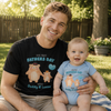 A smiling man and a baby sitting outdoors on a blanket, both wearing matching graphic t-shirts and bodysuits with a cartoon bear design and the text 'Our First Fathers Day 2026 Daddy & Lucas' and 'Our First Brothers Day 2026 Daddy & Lucas'.