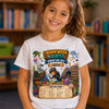 A young girl smiling in a library, wearing a white graphic t-shirt with a colorful 'Book Week Quest Under the Sea' design featuring an underwater scene and a treasure chest chart.