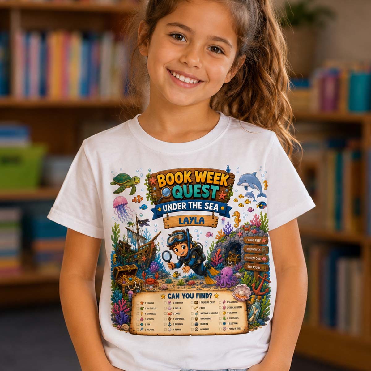 A young girl smiling in a library, wearing a white graphic t-shirt with a colorful 'Book Week Quest Under the Sea' design featuring an underwater scene and a treasure chest chart.