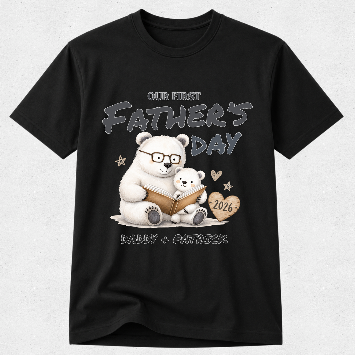 A black short-sleeve crewneck t-shirt featuring a custom Father's Day graphic of a white polar bear and a small cub reading a book, with the text 'OUR FIRST FATHER'S DAY', 'DADDY + PATRICK', and '2026' on a light gray background.