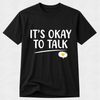 A black short-sleeve crewneck t-shirt with the text 'IT'S OKAY TO TALK' printed in white across the chest, underlined, with a speech bubble containing a small yellow heart below.