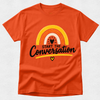 An orange short-sleeve crewneck t-shirt featuring a retro rainbow graphic with the text 'START THE Conversation' in black script and a small yellow heart below, displayed on a light gray textured background.