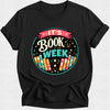 A black short-sleeve crewneck t-shirt featuring a colorful 'IT'S BOOK WEEK' graphic on the chest, displayed against a light gray background.