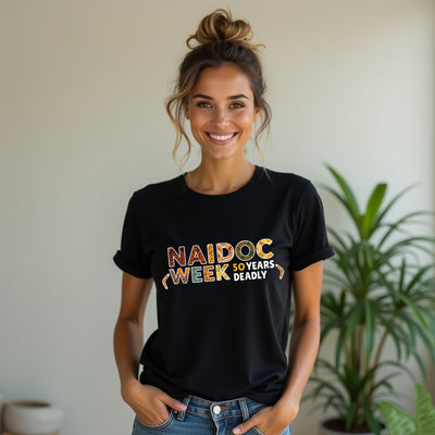 Woman wearing a black t-shirt with 'NAIDOC Week 50 Years Deadly' text, standing indoors with a plant in the background.