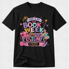 A black short-sleeve crewneck t-shirt featuring a colorful graphic print that reads 'THIS IS MY BOOK WEEK COSTUME!' with illustrations of a pink book, a purple high-heel shoe, a cat-eye sunglasses, and a pink purse on a light gray background.