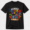 A black short-sleeve crewneck t-shirt featuring a colorful graphic print that reads 'THIS IS MY BOOK WEEK COSTUME!' with cartoon illustrations of a book, superhero cape, dragon, and costume box, shown against a light gray background.