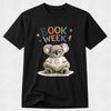 A black short-sleeve crewneck t-shirt featuring a colorful graphic of a cartoon koala wearing glasses and a white 'Teacher Co.' t-shirt, reading a book.