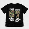 Black t-shirt with camouflage numbers '67' and text 'I was now I'm' on a light gray background