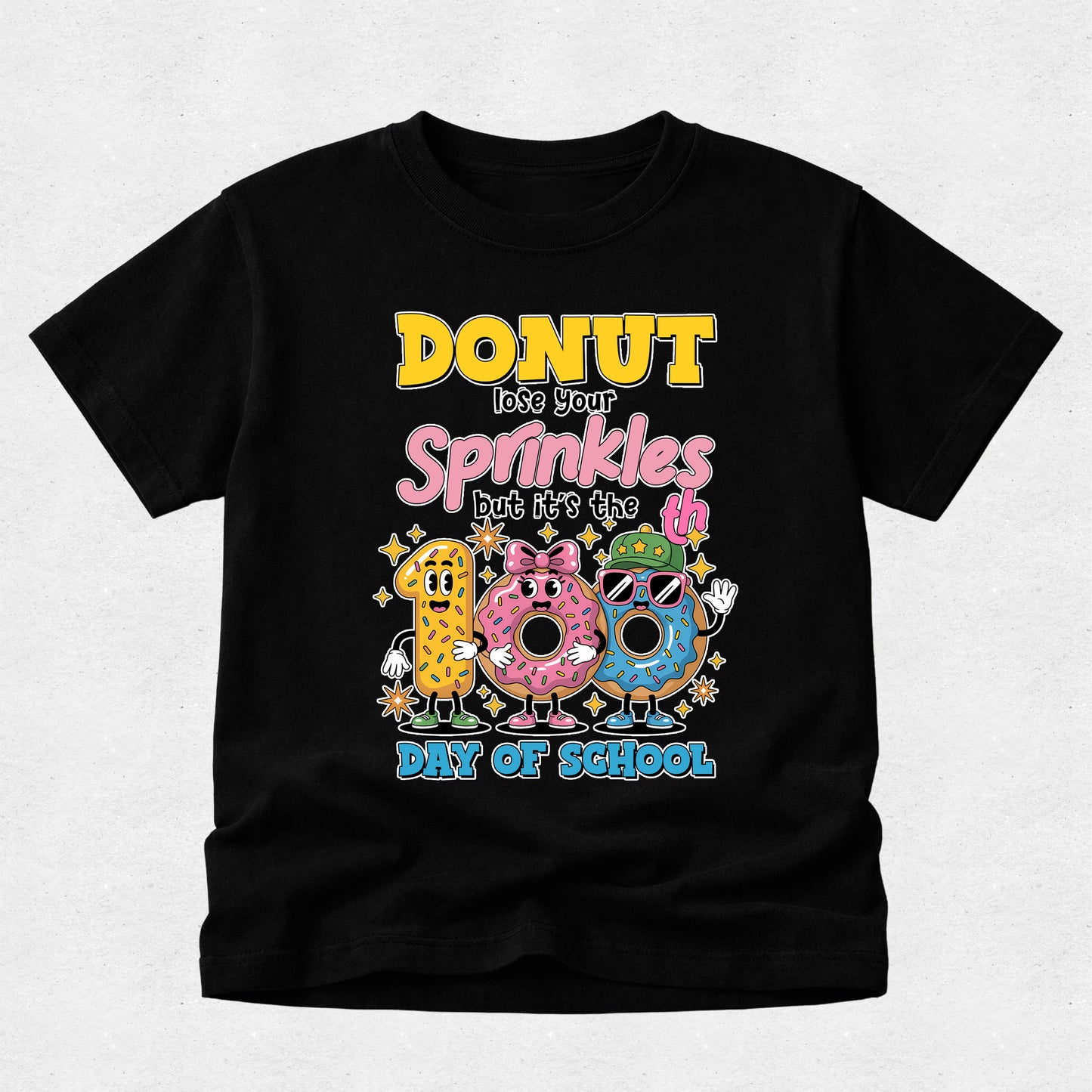 A black short-sleeve t-shirt with a colorful graphic on the front that reads 'DONUT lose your Sprinkles but it's the 100th Day of School' above three cartoon donuts, shown on a light gray background.