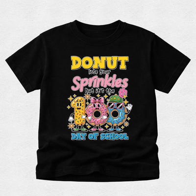 A black short-sleeve t-shirt with a colorful graphic on the front that reads 'DONUT lose your Sprinkles but it's the 100th Day of School' above three cartoon donuts, shown on a light gray background.
