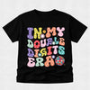 A black short-sleeve t-shirt with a colorful retro graphic that reads 'IN MY DOUBLE DIGITS ERA' in wavy, polka-dotted bubble letters, displayed on a light gray textured background.