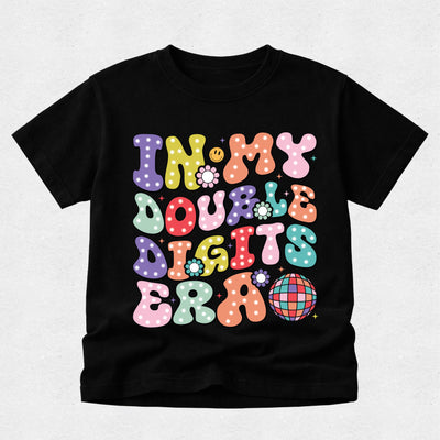 A black short-sleeve t-shirt with a colorful retro graphic that reads 'IN MY DOUBLE DIGITS ERA' in wavy, polka-dotted bubble letters, displayed on a light gray textured background.