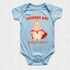 A light blue baby bodysuit with a custom 'Our First Fathers Day Daddy & Julian 2025' graphic featuring two cartoon teddy bears wearing crowns and a red cape, displayed flat against a light gray textured background.