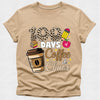 A beige short-sleeve t-shirt with a graphic print that reads '100 DAYS of Coffee & Chaos' in a mix of leopard print, white, brown, and pink fonts, accompanied by illustrations of a coffee cup, books, and a pencil.