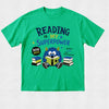 A bright green short-sleeve t-shirt with a colorful graphic on the front. The graphic features the text 'READING is MY SUPERPOWER' in dark blue and purple lettering, along with a cartoon blue monster reading a book, surrounded by stacks of books and a 'BOOK WEEK' badge.