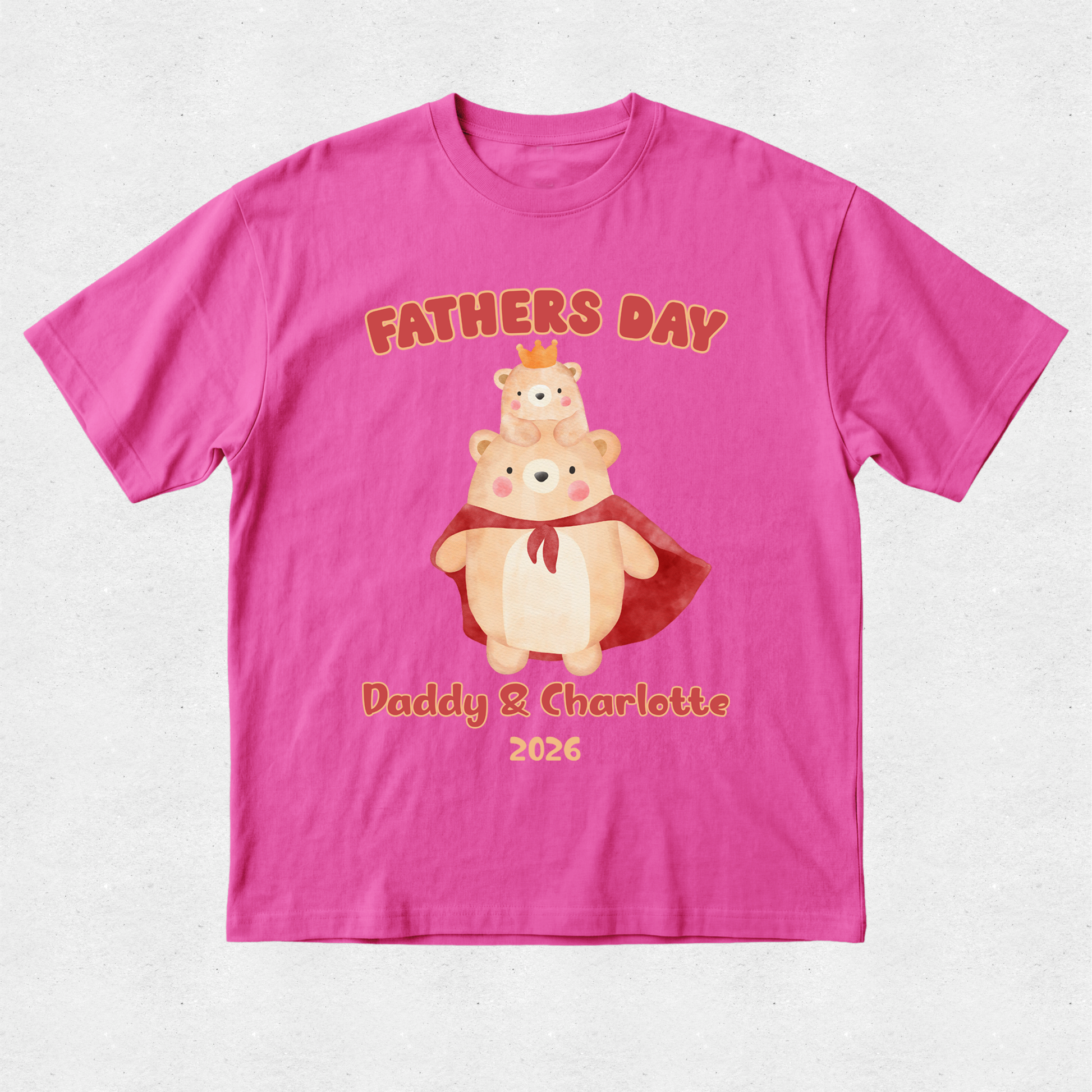 A bright pink short-sleeve t-shirt with a custom graphic on the front. The graphic features two cartoon teddy bears, one wearing a red cape and the other sitting on its shoulders, with the text 'FATHERS DAY' above and 'Daddy & Charlotte 2026' below in a warm orange-brown font.