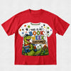 A red short-sleeve crewneck t-shirt featuring a colorful graphic print on the front. The graphic shows a green cartoon dinosaur wearing blue glasses and a red bandana, holding a book, with the text 'THIS IS MY BOOK WEEK COSTUME!' in multicolored letters.