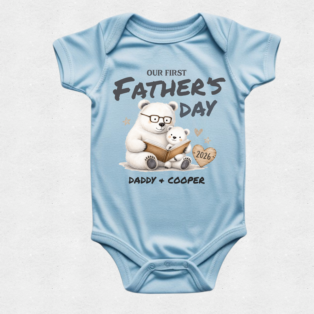 A light blue short-sleeve baby bodysuit with a custom Father's Day graphic. The graphic features a white polar bear wearing glasses and reading a book to a small polar bear, with the text 'OUR FIRST FATHER'S DAY' and 'DADDY + COOPER' along with a heart-shaped tag reading '2026'.