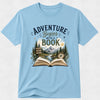 A light blue short-sleeve crewneck t-shirt featuring a graphic print on the front with the text 'ADVENTURE Begins WITH A BOOK' above an illustration of an open book, mountains, pine trees, a river, and a compass.